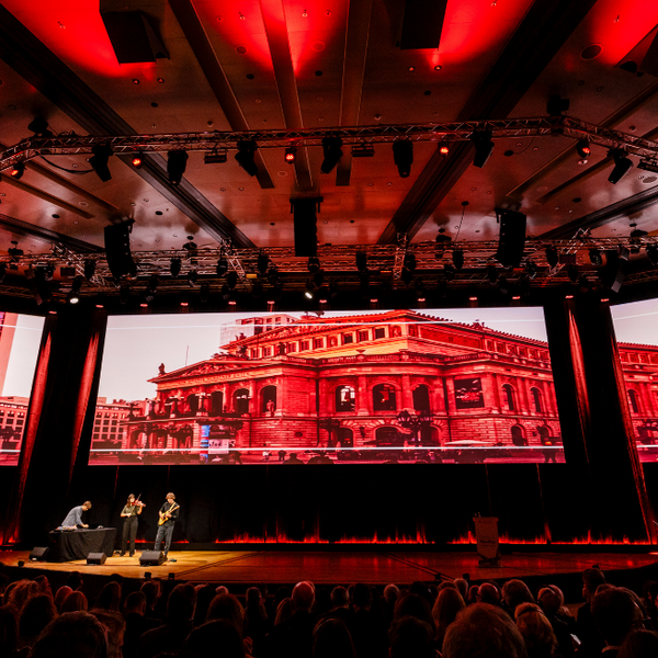 Ein großer, abgedunkelter Saal, beleuchtet durch drei große Leinwände auf denen die Oper Frankfurt zu sehen ist.