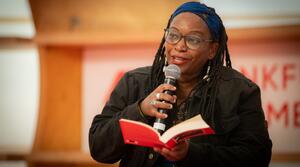 An African author speaks into a microphone while holding an open book in her hand.