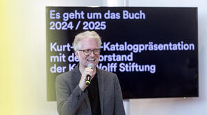 Ein Mann mit Brille steht vor einem Bildschirm und spricht in ein Mikrofon und lächelt dabei. Der Bildschirm zeigt die Aufschrift "Es geht um das Buch 2024/2025 Kurt-Wolff-Katalogpräsentation mit dem Vorstand der Kurt Wolff Stiftung.