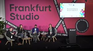 Two women and two men sit next to each other on a stage. The woman on the right holds a microphone and speaks to the audience. There is a lectern to the right of the panellists and a screen in the background shows the title of the event.