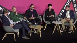 A woman and four men sit next to each other on a stage. The second man from the left holds a microphone and speaks.