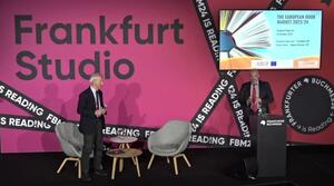 Two chairs and two small tables stand on a stage. To the right is a lectern and behind it a man speaking to the audience. Behind him, a screen shows the title of the event. A man stands in front of the left-hand chair and looks at the other man behind the lectern.