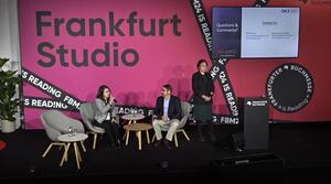 A woman and a man are sitting next to each other on the stage. A woman stands to the right of them. To her right is a lectern. Behind it, a screen shows information about the event. The woman sitting on the left is holding a microphone and speaking to the man next to her.