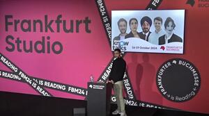 A man stands at a lectern on the stage and speaks to the audience. Behind him, a screen shows two women and three men.