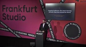 A man stands at a lectern on the stage and speaks to the audience. Behind him, a screen shows the title of the event.