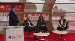 A man stands to the left of the stage at a lectern and looks in the direction of his dialogue partners, a man and two women sitting next to each other on the stage. There are two round tables in the foreground.