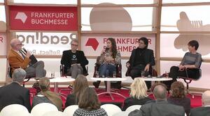 Three men and two women are sitting on a stage. The man on the left holds a microphone in his hand and speaks to the other participants in the dialogue.