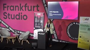 Two chairs and two small tables are placed on a stage to the left. To the right is a roll-up and a lectern at which a woman stands and speaks to the audience. Behind her, a screen shows the title of the event and the name of the speaker. To the right is another roll-up.