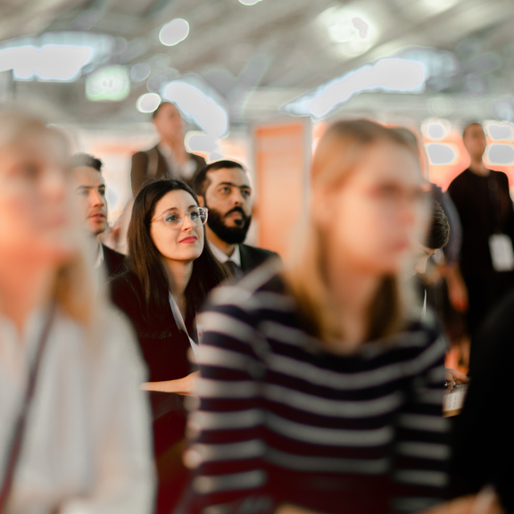 Foto Menschen, die gemeinsam in einer Messehalle sitzend einem Vortrag lauschen. Die Blicke alle in dieselbe Richtung gerichtet. 