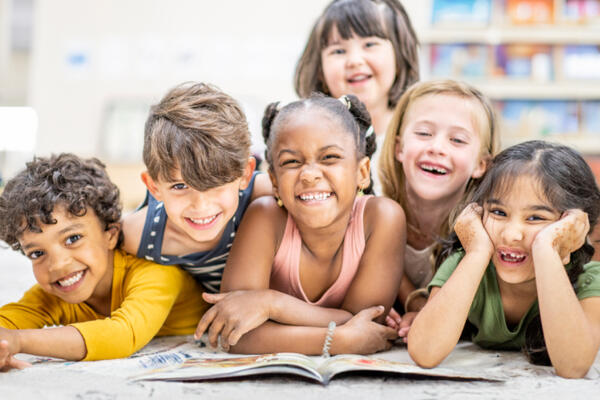 Sechs Kinder liegen grinsend um ein Buch geschart auf einem Teppich.